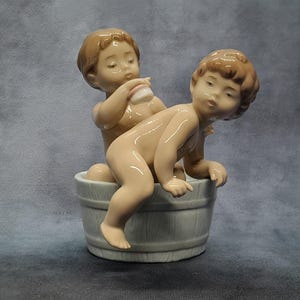 Puede incluir: Figura de porcelana que representa a dos querubines. Uno está sentado en una bañera gris claro, el otro detrás, sosteniendo una esponja. Las figuras tienen cabello castaño claro y están desnudas. La bañera tiene un diseño de barril de madera.