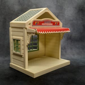 May include: A miniature toy shop with a beige exterior, green-framed window, and a red awning. The sign above the awning reads "GAMES SOUVENIRS". The roof is a textured green. The shop has a small base and is designed to be a playset.