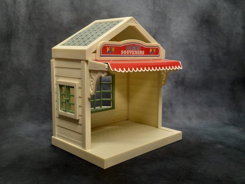 May include: A miniature toy shop with a beige exterior, green-framed window, and a red awning. The sign above the awning reads "GAMES SOUVENIRS". The roof is a textured green. The shop has a small base and is designed to be a playset.