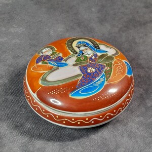 May include: Round, antique ceramic trinket box with a lid. The box is primarily reddish-brown with a white and gold dotted border. The lid features a detailed illustration of figures in shades of orange, blue, green, and white.