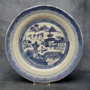 May include: A white ceramic plate with a blue and white design of a Chinese landscape with pagodas and trees. The plate has a scalloped edge.
