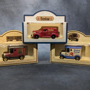 Può includere: Tre camion giocattolo vintage in scatole espositive. I camion sono rossi, blu e amaranto con ruote dorate. Le scatole presentano i loghi "Days Gone" e "Hamleys".