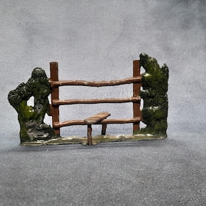 Puede incluir: Una valla rústica en miniatura con un pequeño banco, flanqueada por dos árboles verdes y musgosos texturizados. La valla es marrón con tres rieles horizontales. La escena está sobre una superficie gris.