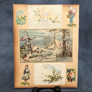 May include: Vintage Christmas card with a central winter scene of a snow-covered cottage. The card features illustrations of children, flowers, and doves, with the text "A joyful Christmas" and "A happy and joyful Christmas".