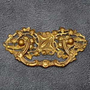 May include: Ornate gold-colored drawer pull with a floral and leaf design. The pull features a central square element with a leaf pattern, flanked by curved handles and decorative accents. A vintage-style hardware piece.