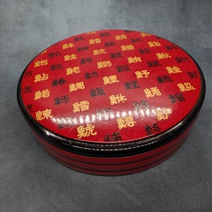 Op de afbeelding: Een ronde, rode lakdoos met een glanzende afwerking, voorzien van een herhalend patroon van zwarte en gouden Chinese karakters op het deksel. De zijkanten van de doos zijn ook rood met dunne, horizontale zwarte lijnen rondom.