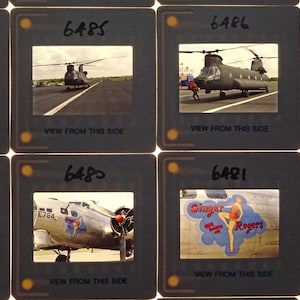 May include: A collection of eight vintage 35mm slides featuring various aircraft, including a helicopter, a bomber, and a plane with a "Ginger Rogers" pinup girl design. The slides are labelled with numbers and the phrase "View from this side".