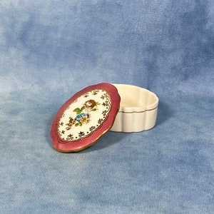 Op de afbeelding: Een klein, ovaal sieradendoosje met een roze deksel en een witte basis. Het deksel heeft een bloemmotief met gouden accenten. Het doosje is open en het deksel ligt naast de basis.