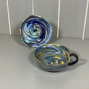 Può includere: Un set da tè composto da tazza e piattino con un motivo a spirale blu e giallo. La tazza ha un manico nero.