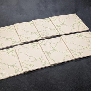 Puede incluir: Un conjunto de siete azulejos de cerámica rectangulares dispuestos en dos filas. Cada azulejo presenta una superficie brillante de color blanco roto con un patrón de mármol veteado en verde claro. Los azulejos tienen superficies lisas.