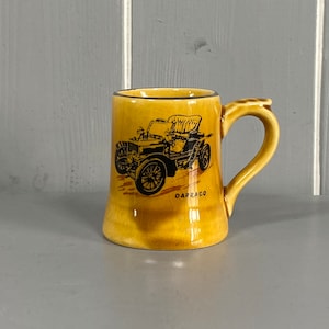 Puede incluir: Una taza de cerámica amarilla con una imagen en blanco y negro de un coche antiguo. El coche es un Darracq, una marca francesa de automóviles. La taza tiene un asa y un borde negro.