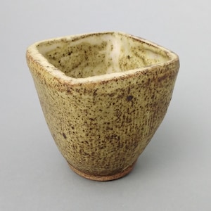 Può includere: Una piccola ciotola quadrata in ceramica con una glassa verde macchiata. La ciotola ha un fondo leggermente arrotondato e una superficie ruvida e testurizzata.