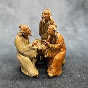 Op de afbeelding: Een keramieken beeldje dat drie oude mannen in gewaden afbeeldt. De figuren zijn in tinten beige en oranje, met gedetailleerde gelaatstrekken en lange baarden. Ze lijken verzameld te zijn rond een tafel.