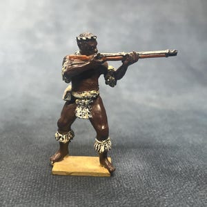 May include: A miniature figurine of a person holding a long rifle, painted in brown and tan tones. The figure has decorative elements around the waist, arms, and legs. The figurine stands on a small, rectangular base.