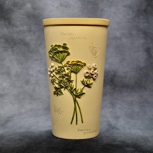 May include: A tall, cream-colored ceramic vase with embossed floral designs and botanical text. The vase features a bouquet of green and yellow flowers with white accents. The text reads "Torilis japonica" and "Anethum graveolens".