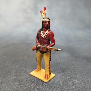 May include: A painted metal figurine of a Native American man holding a rifle. The figure is painted with red skin, tan trousers with fringe, and a feathered headdress. The figure stands on a small, rectangular base.