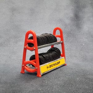 K&ouml;nnte beinhalten: Ein kleiner roter Reifenst&auml;nder mit zwei Stapeln schwarzer Reifen. Der St&auml;nder hat ein gelbes Schild mit dem Dunlop-Logo. Die Reifen bestehen aus einem gummiartigen Material und sind ordentlich auf den Metallregalen gestapelt.