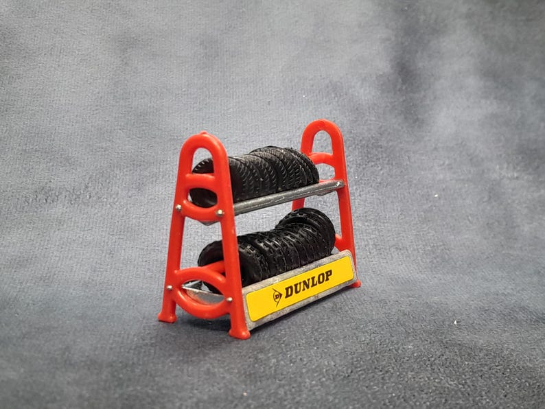 K&ouml;nnte beinhalten: Ein kleiner roter Reifenst&auml;nder mit zwei Stapeln schwarzer Reifen. Der St&auml;nder hat ein gelbes Schild mit dem Dunlop-Logo. Die Reifen bestehen aus einem gummiartigen Material und sind ordentlich auf den Metallregalen gestapelt.
