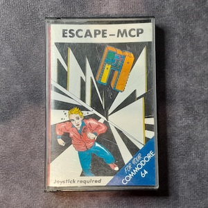 May include: A retro Commodore 64 game cassette labelled "ESCAPE-MCP." The cover art shows a vintage illustration of a running figure, a computer, and geometric shapes. The text "Joystick required" is also visible.