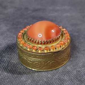 Puede incluir: Pequeña y redonda caja de baratijas antigua con una tapa de piedra color coral. La caja es dorada con intrincados diseños florales y un anillo de pequeñas piedras anaranjadas.