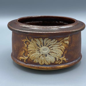 Op de afbeelding: Een bruine keramische plantenpot met een reliëf wit bloemenontwerp. De pot heeft een afgeronde vorm en een licht uitlopende rand.