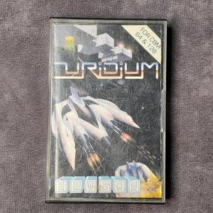 Puede incluir: Un cartucho de videojuego Uridium vintage para Commodore 64 y 128. La portada presenta un diseño de nave espacial futurista con el título del juego en letras doradas. El logotipo de Hewson está en la parte inferior.