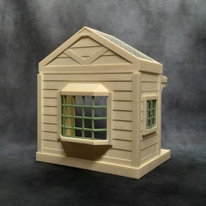 May include: A miniature beige house model with a gabled roof and a small window with green grid panes. The house has horizontal siding and a small window on the side. The roof is a darker shade of green.