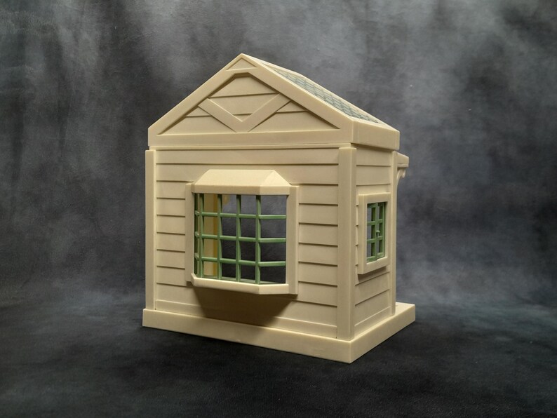 May include: A miniature beige house model with a gabled roof and a small window with green grid panes. The house has horizontal siding and a small window on the side. The roof is a darker shade of green.