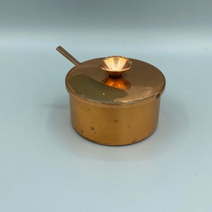 Könnte beinhalten: Eine kleine, runde Zuckerdose aus Kupfer mit Deckel und kleinem Löffel. Die Dose hat eine glänzende, metallische Oberfläche.