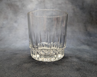 May include: A clear, cylindrical drinking glass with a patterned base and vertical lines etched into the sides. The glass is empty and sits on a dark surface, showcasing its transparent design.