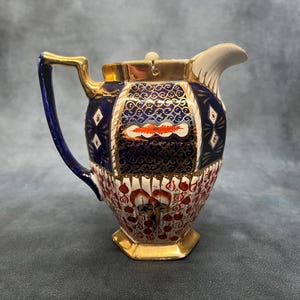 Könnte beinhalten: Eine antike Keramikkrug mit goldenem Griff und Sockel. Der Krug hat einen marineblauen Hintergrund mit goldenen Filigranarbeiten und rot-weißen Blumenmustern. Der Ausguss ist weiß.