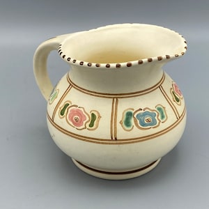Może przedstawiać: Biały dzbanek ceramiczny z brązowym uchwytem i kwiatowym wzorem. Dzbanek ma brązowy, zielony, różowy i niebieski wzór kwiatowy.
