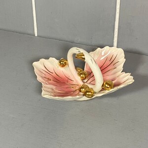 May include: A white ceramic dish shaped like two leaves with a gold handle. The dish is decorated with pink and gold accents and has gold balls around the handle.