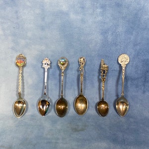 Peut inclure: Sept cuillères à café souvenirs en argent avec des motifs différents. Chaque cuillère a une petite image colorée sur le manche.