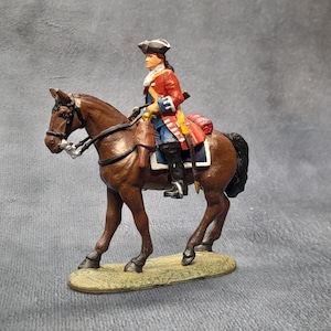 May include: A miniature figurine of a soldier riding a brown horse. The soldier wears a red coat, blue pants, and a black hat. The horse has a black mane and tail, with a saddle and bridle. The figurine is on a small, green base.
