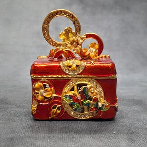 May include: A red, rectangular trinket box with gold-colored floral and jewel accents. The box has a hinged lid with a circular handle. The sides feature ornate, circular designs with colorful floral details.