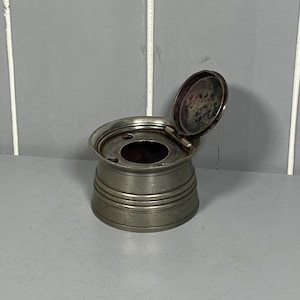 May include: A silver inkwell with a hinged lid. The lid is open and the inkwell has a round opening in the top.