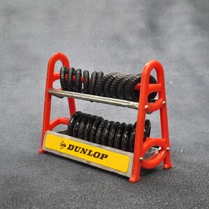 K&ouml;nnte beinhalten: Ein rotes Miniatur-Reifenregal mit mehreren schwarzen Reifen. Das Regal hat zwei Ebenen und ein gelbes Schild mit dem Dunlop-Logo. Die Reifen sind ordentlich gestapelt, was auf ein Modell oder Spielzeug hindeutet.
