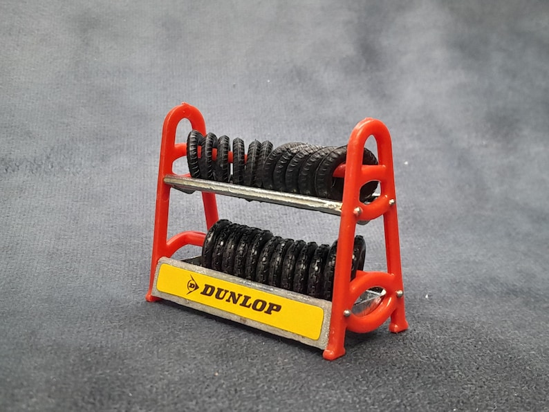 K&ouml;nnte beinhalten: Ein rotes Miniatur-Reifenregal mit mehreren schwarzen Reifen. Das Regal hat zwei Ebenen und ein gelbes Schild mit dem Dunlop-Logo. Die Reifen sind ordentlich gestapelt, was auf ein Modell oder Spielzeug hindeutet.