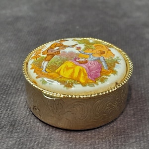May include: A small, round, gold-coloured trinket box with a detailed lid. The lid features a painted scene of two figures in a garden setting, with a yellow and pink colour palette. The box has a decorative, embossed pattern.