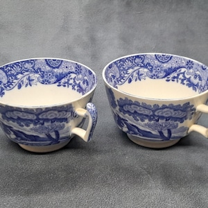 May include: Two cream-colored teacups with blue floral and landscape patterns. The rims and handles are detailed with blue designs. The cups are placed side by side, showcasing their matching aesthetic.
