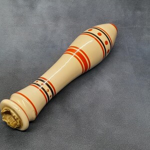 May include: A decorative bottle stopper with a cream-coloured body and red, black, and orange accents. The stopper has a rounded top and a cork base. The design includes red stripes and black dotted bands.