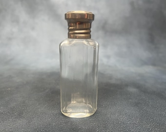 May include: A clear glass perfume bottle with a gold-coloured cap. The bottle has a rectangular shape with a slightly rounded base. The cap is a darker gold colour and has a ribbed design. The bottle is empty and appears to be vintage.