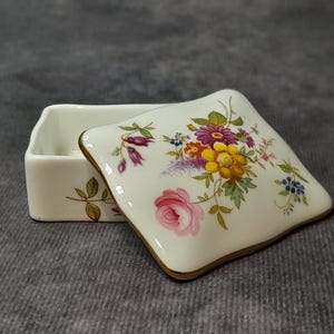 Puede incluir: Una pequeña caja de porcelana blanca abierta con un diseño floral en la tapa. La tapa presenta una rosa rosa, flores moradas, amarillas y azules con hojas verdes. La caja tiene un borde dorado.