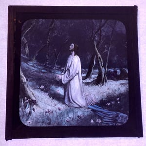 May include: An antique slide featuring a religious painting. The artwork depicts a figure in a white robe kneeling in a forest, with hands clasped in prayer. The scene is set in a dark, wooded area with a stream.