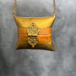 Pode incluir: Um pequeno pendente de latão em forma de almofada com um fecho decorativo e uma faixa de cobre. O pendente pende de uma corrente delicada. O fecho e a faixa apresentam desenhos intrincados e ornamentados. A estética geral é vintage e antiga.