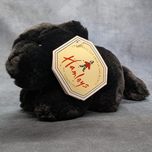 May include: A plush black animal toy with soft fur, shown in a lying position. Attached to the toy is a cream-colored, octagonal paper tag featuring the red Hamleys logo, a small illustration of a toy soldier, and the text The Finest Toy Shop In The World.