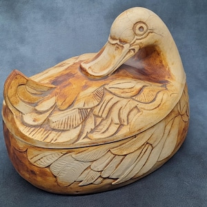 May include: A duck-shaped box with a lid, crafted from light brown wood with darker brown accents. The box features carved feather details. The duck's head is turned to the side. The box is set against a blue backdrop.