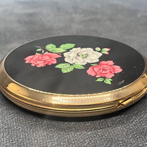 Op de afbeelding: Een vintage compacte spiegel met een zwarte emaille bovenkant. De bovenkant is versierd met een bloemenontwerp met roze en witte rozen en groene bladeren. De compact heeft een goudkleurig metalen frame en een scharniersluiting.
