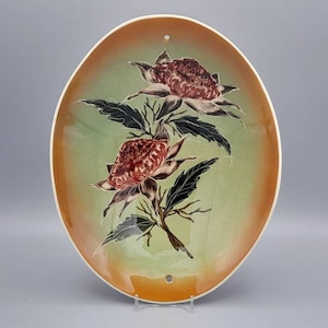 May include: A ceramic wall plate with a green and orange gradient background. The plate features a painted design of two red and white flowers with black stems and leaves.